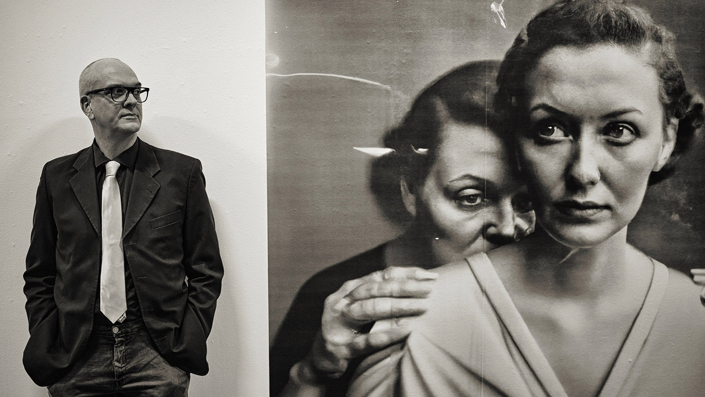 Black and white photograph of a man in a suit with a white tie standing next to a large vintage-style artwork. The man, wearing glasses and looking to the left, stands against a plain wall, while the artwork beside him depicts two feminine humans, one in the foreground with a serious expression, and another seemingly whispering or leaning on the sholder of the other from behind.